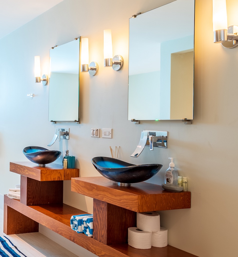 Luxury Bonaire villa spa bathroom with a custom wood double vanity, blue glass vessel sinks, and modern chrome waterfall faucets.